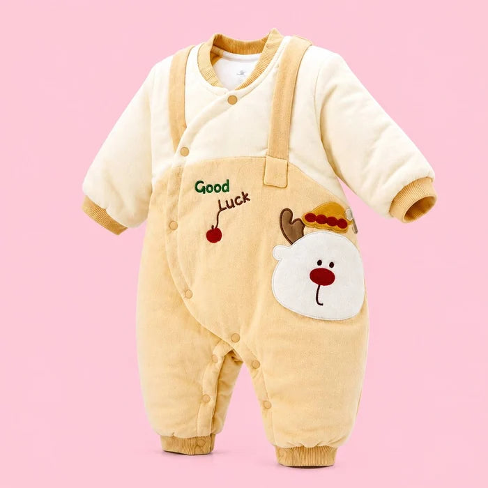 A baby's long-sleeve corduroy jumpsuit in cream and light tan, featuring a white bear face patch on the front pocket, a small red cherry with 'Good Luck' text, and adjustable snap closures, set against a solid pink background.