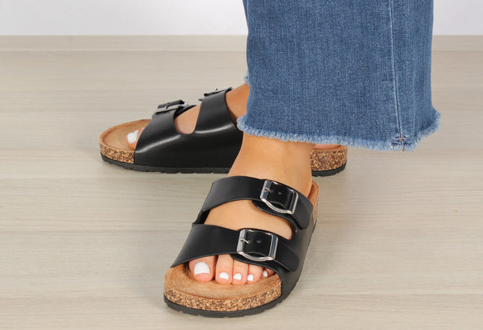 A foot wearing a double strap sandal with silver buckles, the sandal is black and white, the buckle is silver, the foot is in a relaxed position.