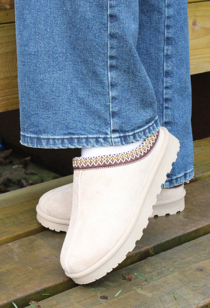 A person wearing slip-on shoes with soft faux fur lining and embroidered trim, in a color best described as taupe, paired with blue jeans.