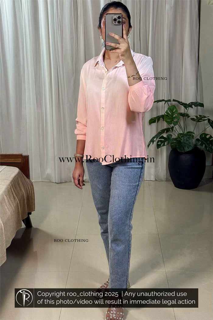 Person wearing a light pink shirt and blue jeans taking a mirror selfie in a room with a bed and plant.