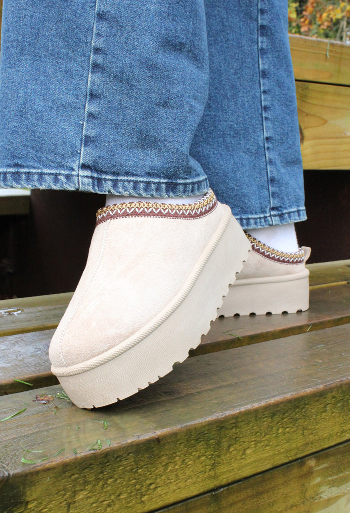 A person wearing blue jeans and white platform slip-on boots with embroidered trim sitting on a wooden bench.