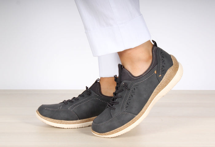 A pair of navy slip-on trainers with elastic laces and laser cut detailing, worn by a person standing on a light wooden floor.
