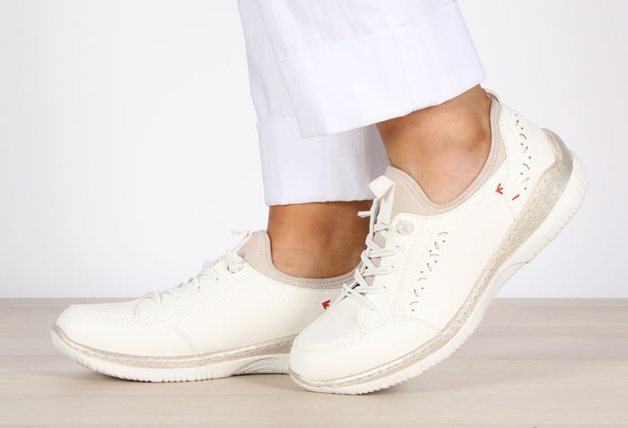 A pair of white slip-on trainers with elastic laces and laser cut detailing, worn by a person standing on a light wooden floor.