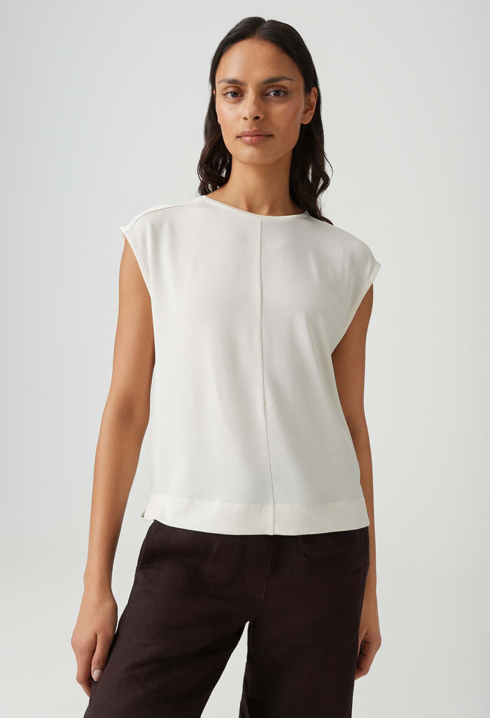 Woman wearing a white sleeveless top and dark pants on a light gray background