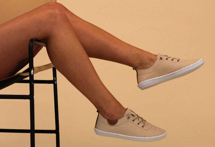 A person seated with their feet hanging off the chair, wearing beige leather plimsolls with lace-up closure.