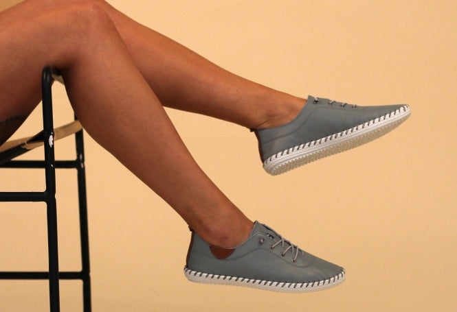 A person seated with their feet hanging off the chair, wearing blue leather plimsolls with lace-up closure.