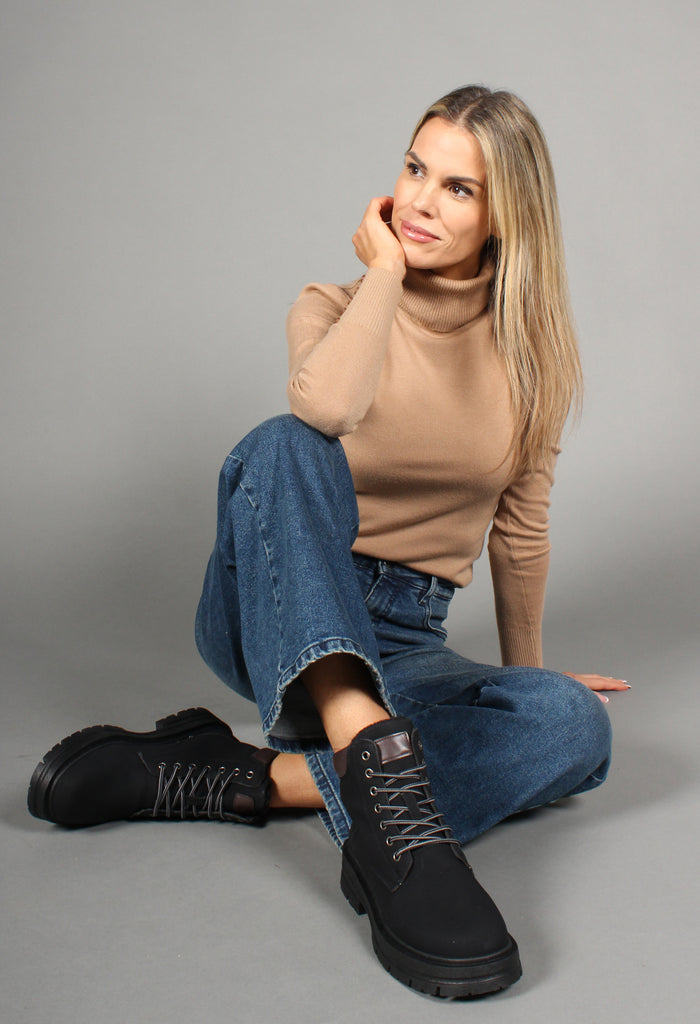 A woman sitting on a grey background wearing a beige turtleneck sweater, blue jeans, and black 'Working Style Boots' with a round toe, lace-up front, faux fur lining, and a block heel.